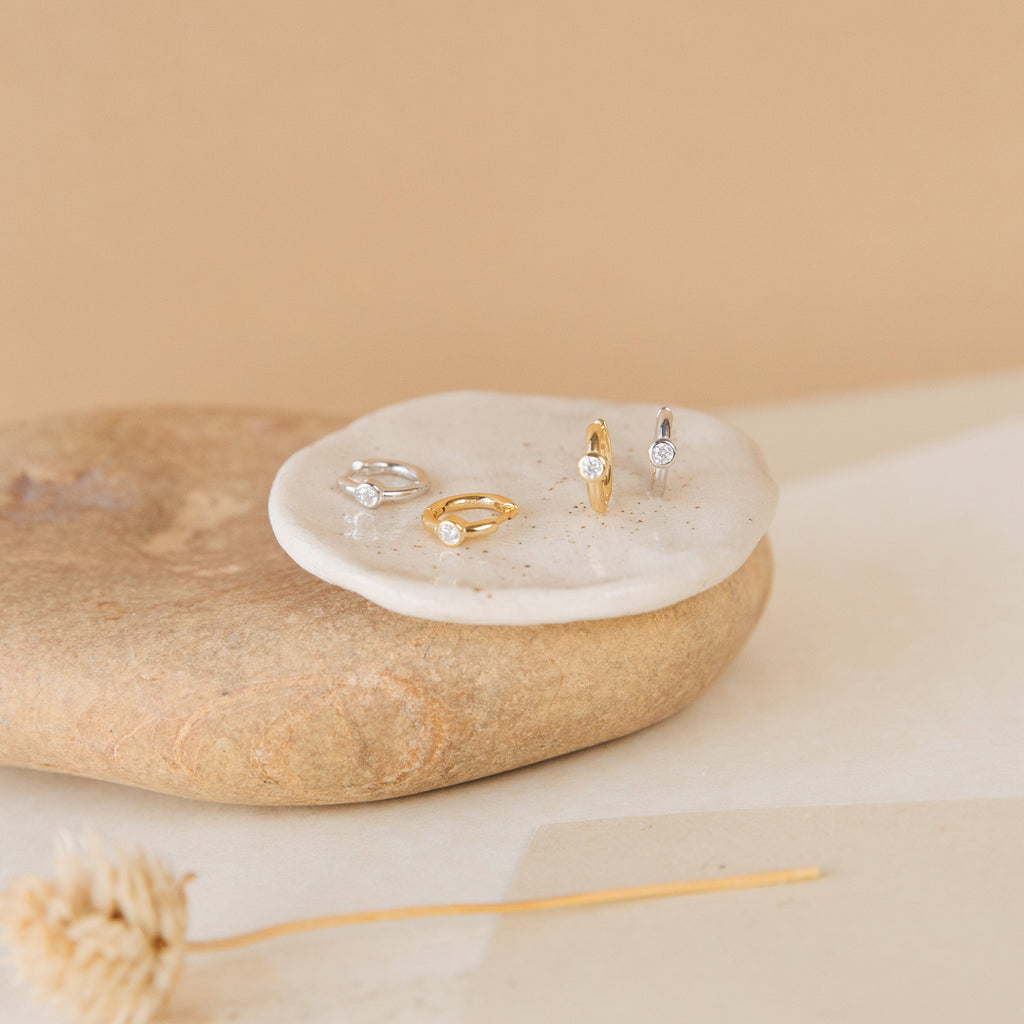 Aela Huggies earrings and delicate gold and silver rings with small gems are displayed on stone and ceramic dishes against a beige background.